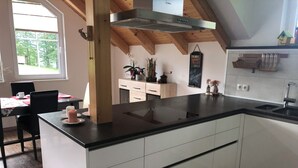 Une cuisine moderne dotée d’un îlot central, de meubles blancs et d’un plan de travail noir. Une fenêtre laisse entrer la lumière naturelle, et on peut apercevoir une structure en bois au plafond.