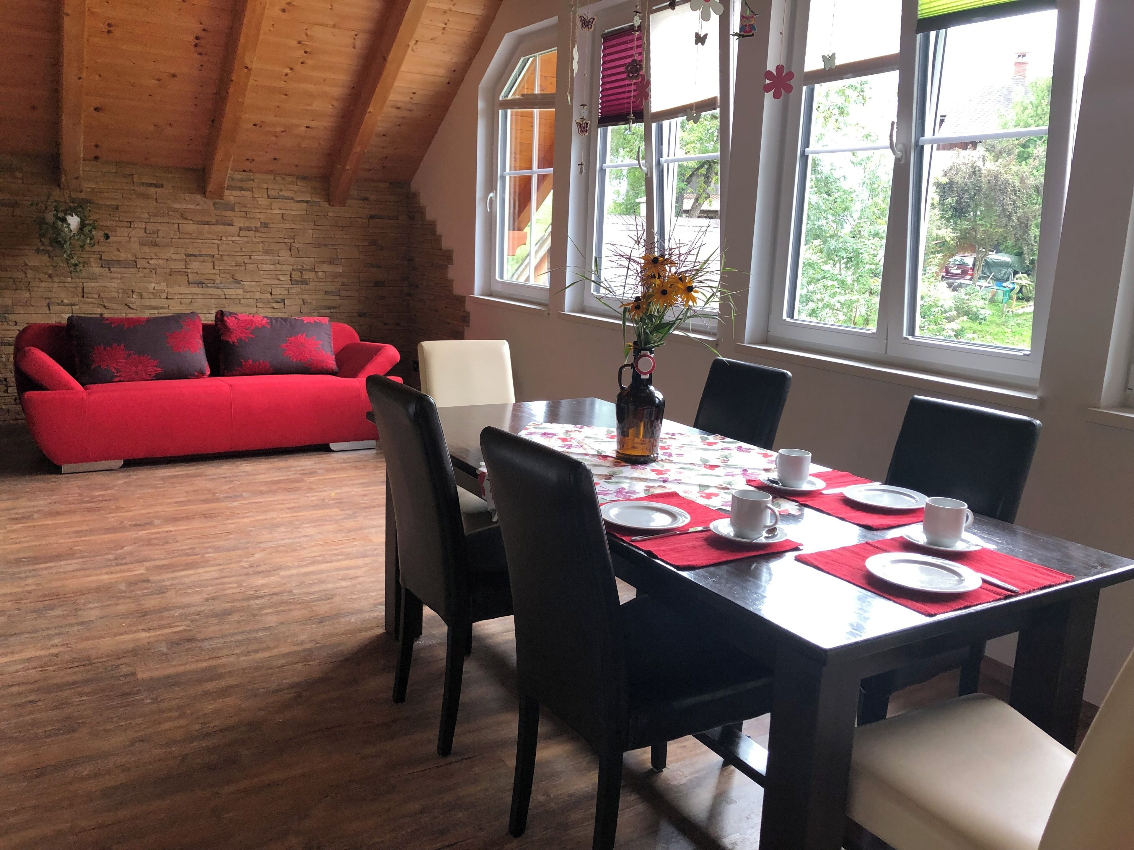 A dining area with a wooden table set for four, surrounded by chairs, and a red sofa in the background.
