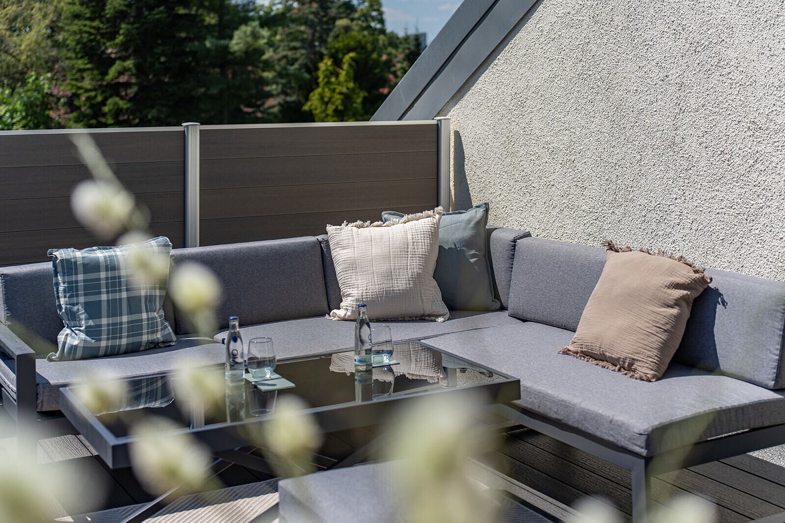 Ein Außenbereich mit Tisch mit Glasplatte, grauem Sofa mit Polstern und verschiedenen Kissen.