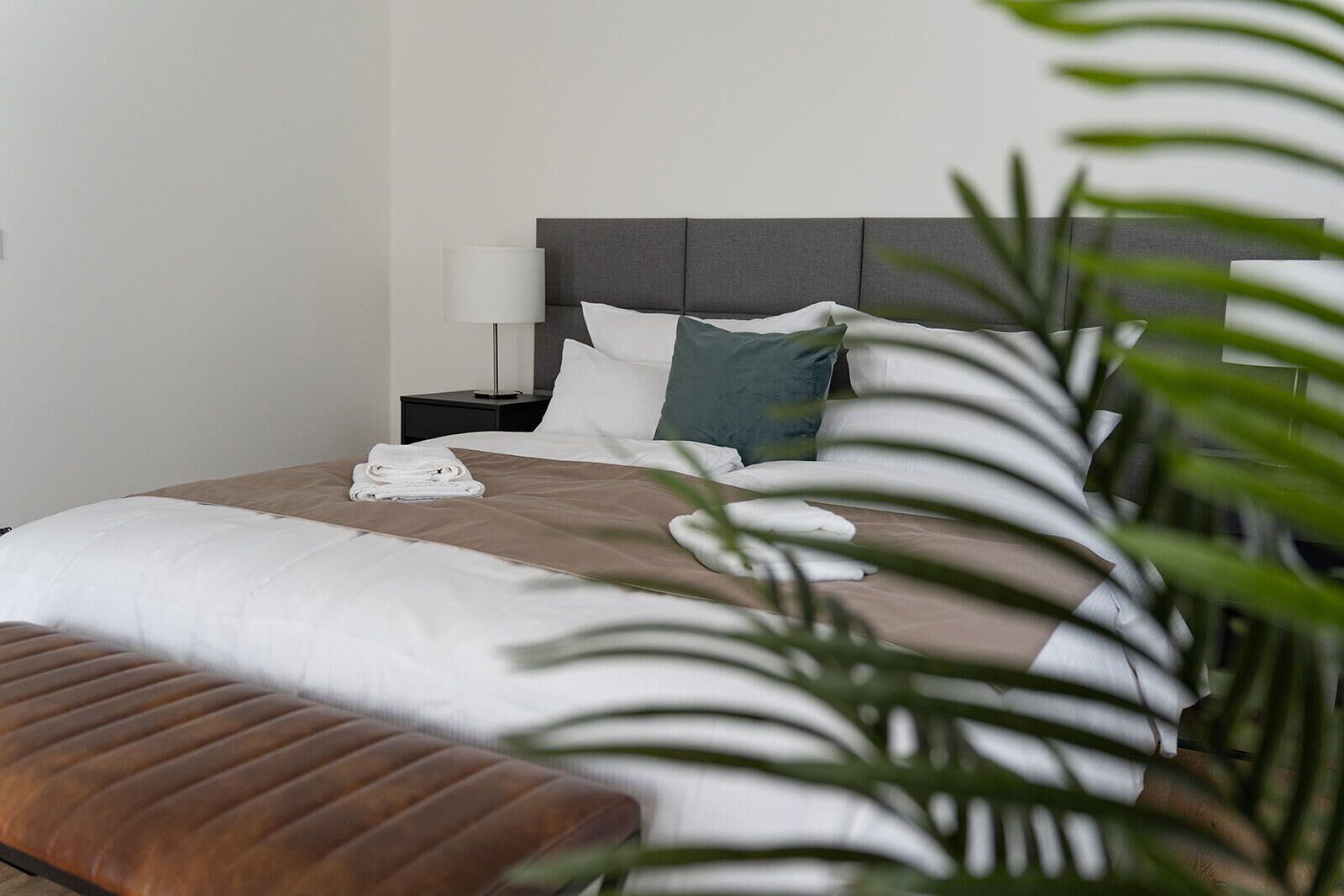 Ein ordentlich bezogenes Bett mit einer braunen Tagesdecke, weißen Kissen und einem gefalteten Handtuch.