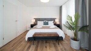 A modern hotel room with a large bed, bedside tables, a bench, a potted plant, and a ceiling light.