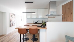 A modern kitchen with white cabinets, a wooden countertop, and a stainless steel range hood. There are two stools at the island and a painting on the wall.