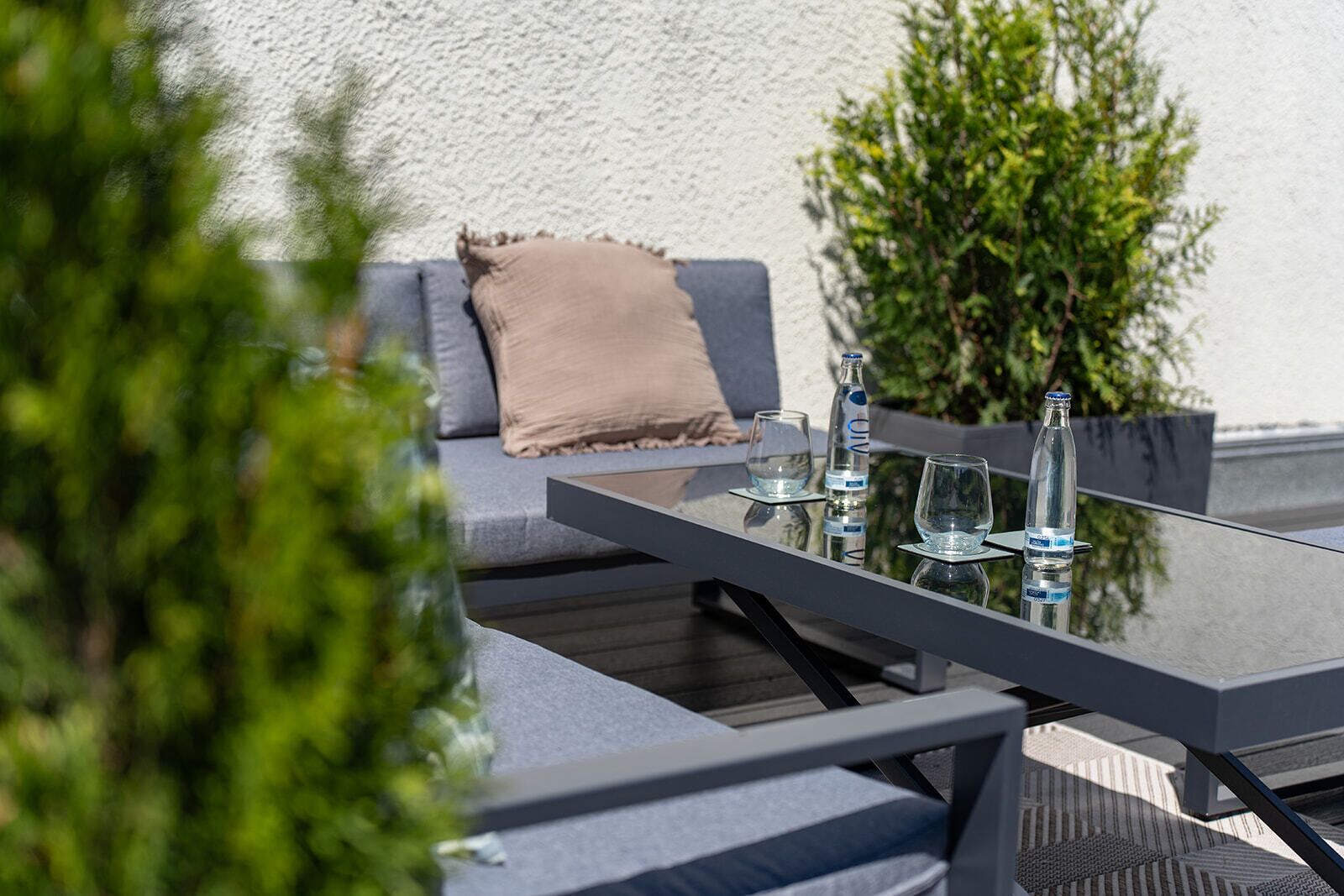 A modern outdoor seating area with a glass table, two chairs, a sofa with cushions, and a bottle of water.