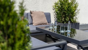 A modern outdoor seating area with a glass table, two chairs, a sofa with cushions, and a bottle of water.