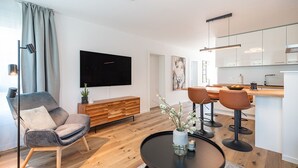 A modern living room with a flat-screen TV, a wooden coffee table, a grey sofa, and a dining area with a kitchen in the background.