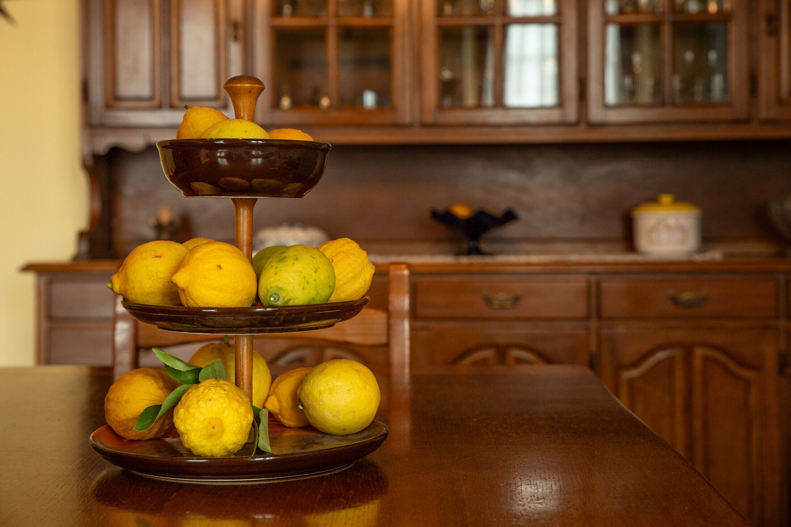 A three-tiered fruit stand with lemons and limes on a wooden table in a kitchen with wooden cabinets.