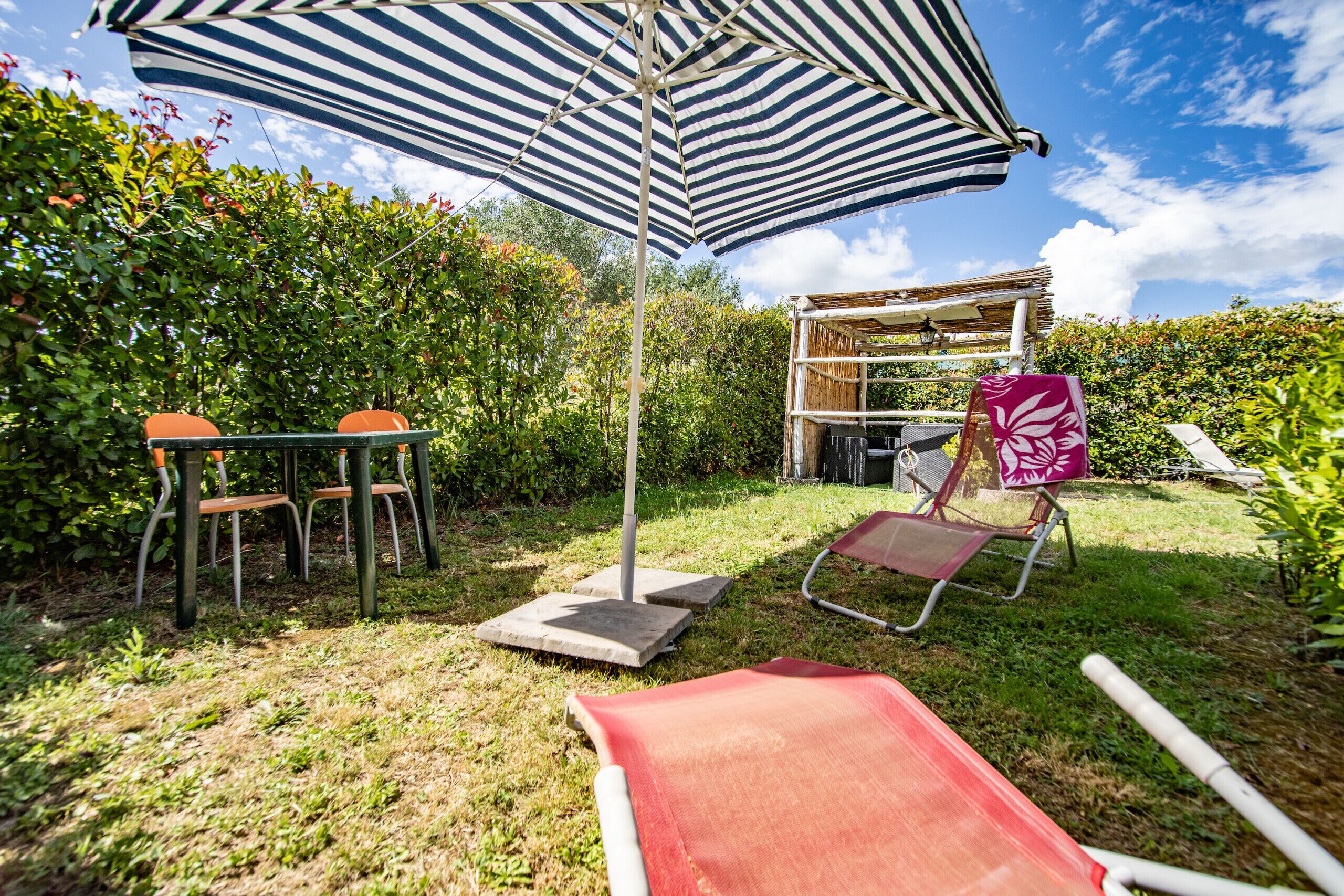 An outdoor patio with a striped umbrella, a table with chairs, a lounge chair, and a small wooden structure.