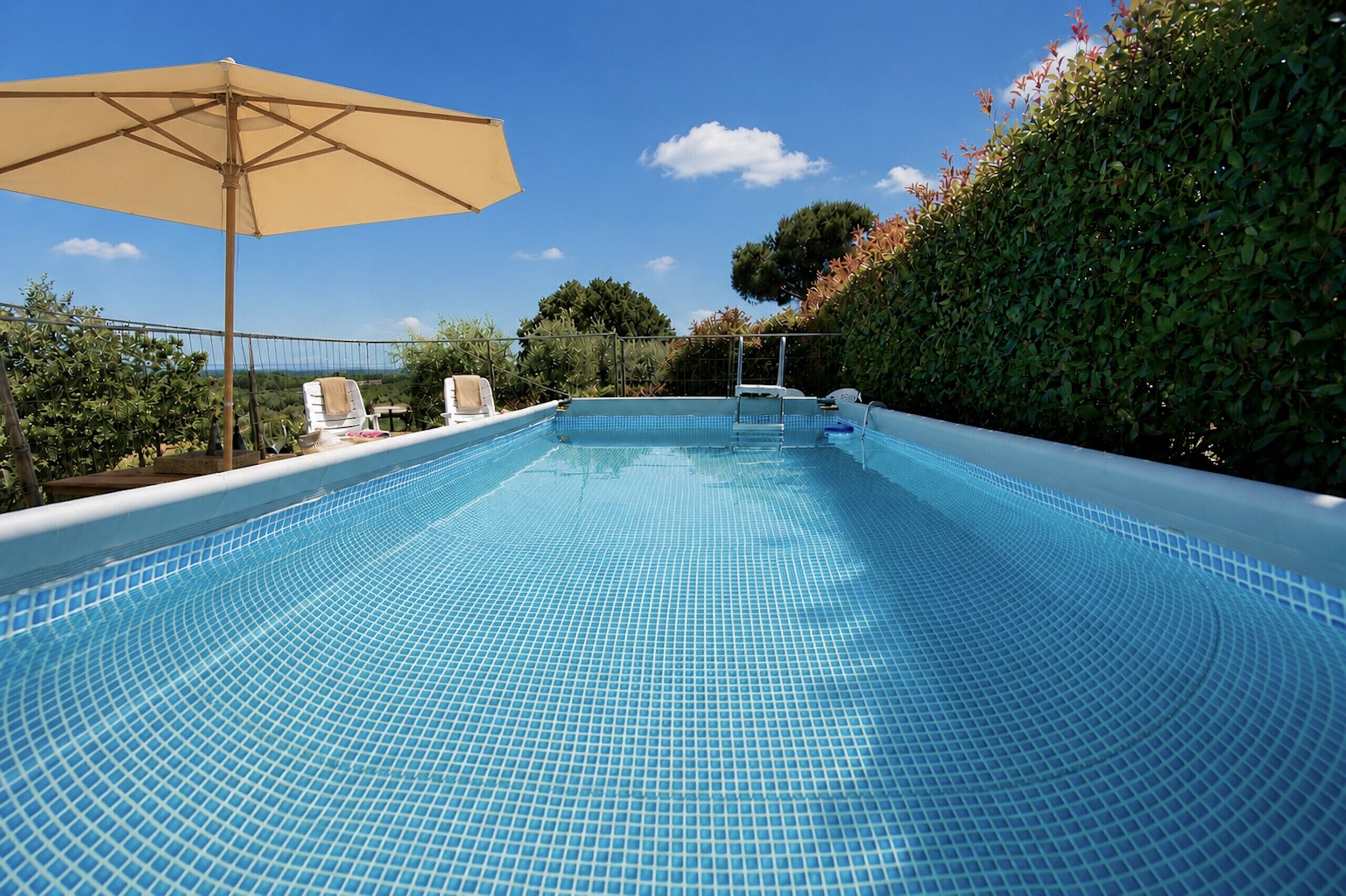 Ein Swimmingpool mit Fliesenboden, umgeben von einer Hecke, und ein Liegestuhl unter einem Sonnenschirm.