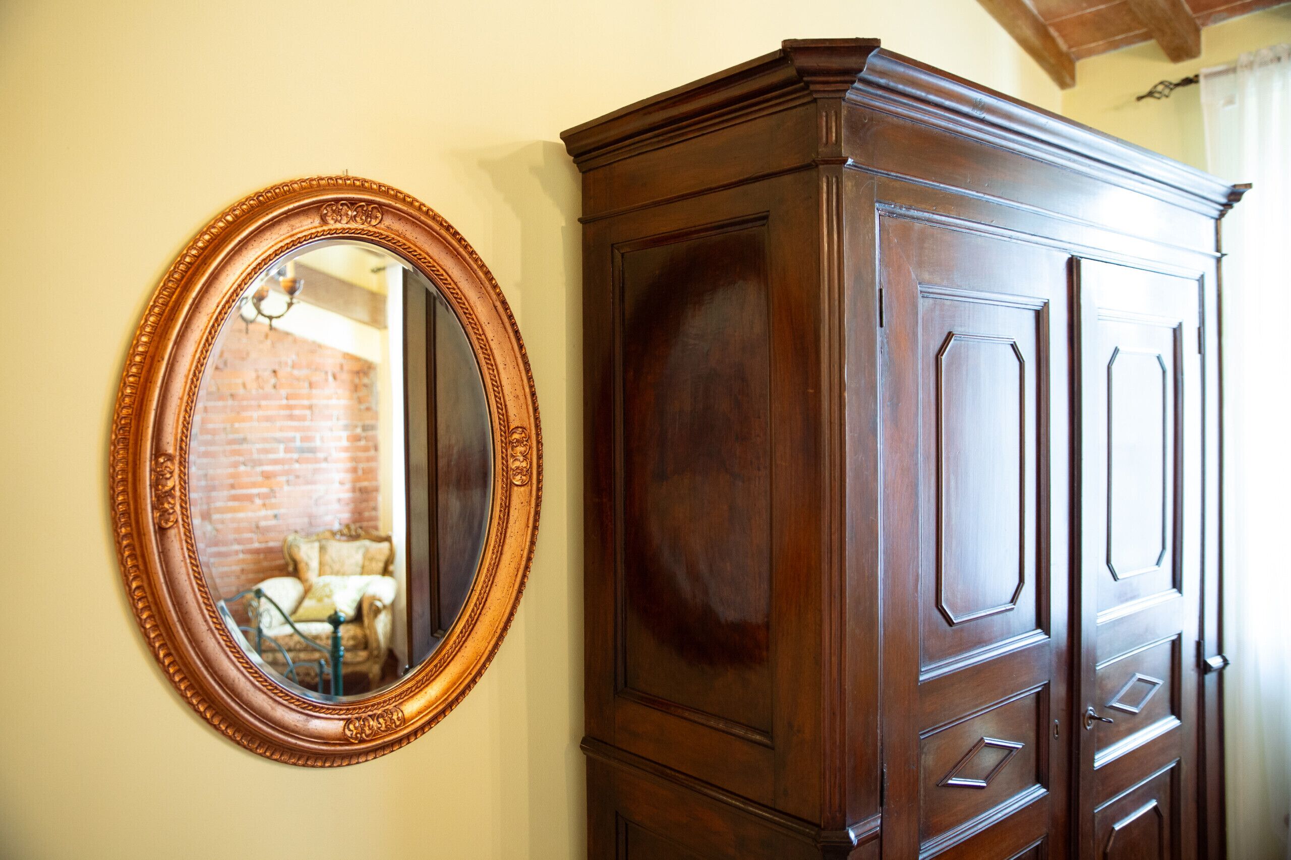 A wooden wardrobe with multiple doors and drawers, a mirror with an ornate frame, and a brick wall in the background.