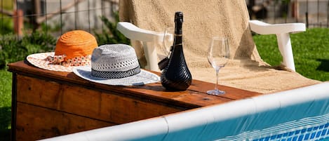 A poolside setting with a wooden table, a bottle, a glass, and two hats.