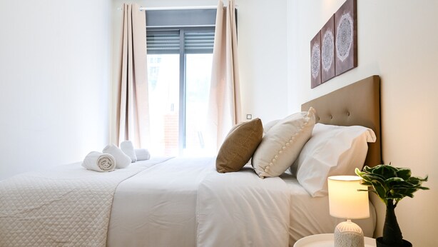 Ein ordentlich bezogenes Bett mit einer gesteppten Decke, einer Nachttischlampe und einem Blumentopf auf einem Nachttisch.