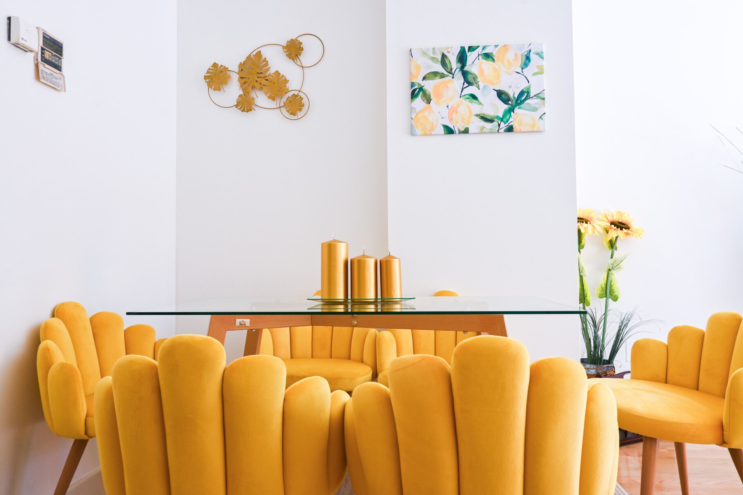 Un coin repas avec une table à plateau en verre, des chaises recouvertes d’un tissu jaune et un vase rempli de fleurs jaunes.