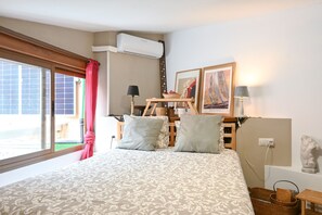 A bedroom with a large bed, a window with curtains, an air conditioning unit, a wooden side table with a lamp, and framed pictures on the wall. - Cozy studio with pool in Ciudad Lineal (Madrid)