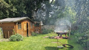 A wooden cabin with a chimney, a barbecue grill, and a small shed in a grassy backyard surrounded by trees.