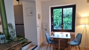 A dining area with a wooden table and two chairs, a window with a view of greenery, and a lamp on the right.