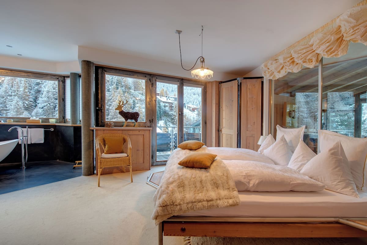 A bedroom with a large bed, a wooden chair, a window with a view of snow-covered trees, and a bathroom with a bathtub and a sink.