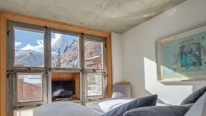 A bedroom with a large window, a bed, a television, and a framed picture on the wall. - Backstage Hotel Serviced Apartments (Zermatt)