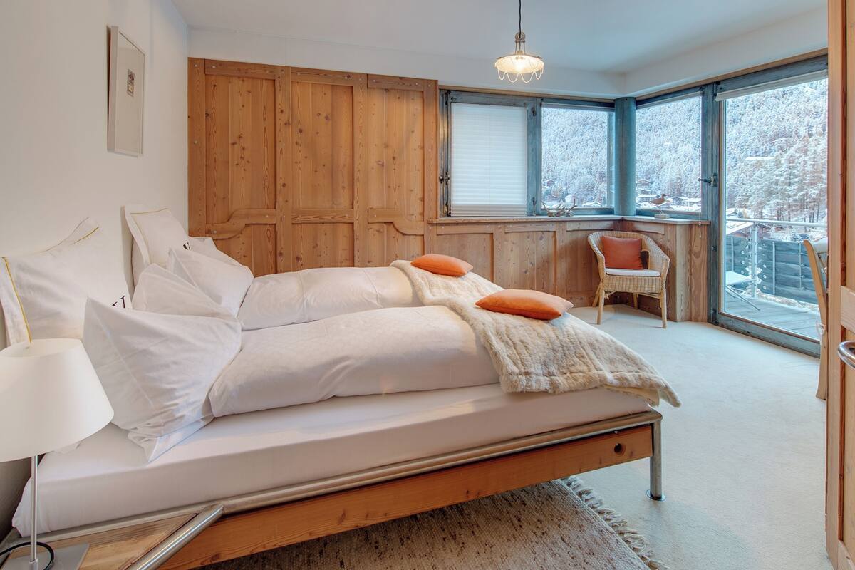 A bedroom with a wooden bed, white bedding, a window with a view of snow-covered trees, and a wicker chair by the window.