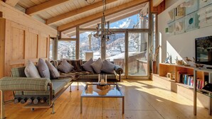 A spacious living room with a wooden ceiling, a large window offering a mountain view, a sofa with cushions, and a glass coffee table. - Backstage Hotel Serviced Apartments (Zermatt)