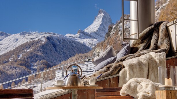A wooden deck with a kettle, teacup, and saucer, pillows, and a fur throw. A mountain with a snow-covered peak is visible in the background. - Backstage Hotel Serviced Apartments (Zermatt)