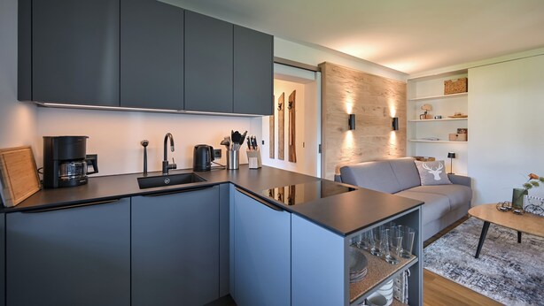 Een moderne keuken met grijze kastjes, een koffiezetapparaat en een spoelbak. Er is een grijze bank en een kleine tafel met een vaas in de woonkamer.