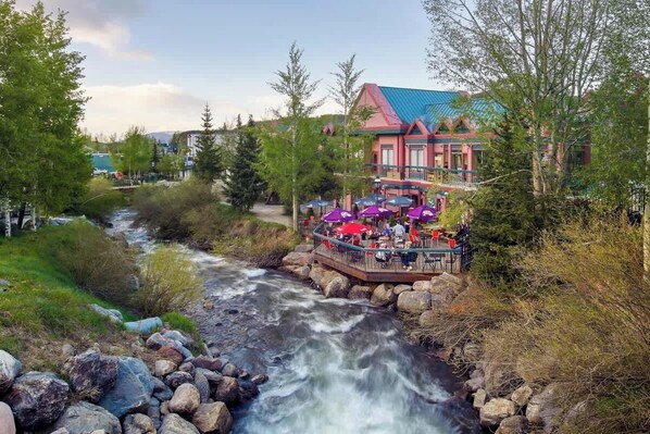 Un restaurant situé au bord de la rivière, avec une terrasse offrant une vue sur le cours d’eau, entouré d’arbres et de bâtiments.