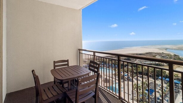 Een balkon met een houten tafel en stoelen, met uitzicht op een strand en zwembad.