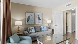 A living room with a sofa, a glass coffee table, and two wall-mounted lamps. There are framed pictures of birds on the wall. - Beachfront Serenity 1 Bedroom 2 Bathroom Escape (North Myrtle Beach)