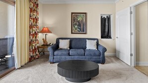 Ein Wohnzimmer mit einem blauen Sofa, einem runden schwarzen Hocker, einer Lampe und einem gerahmten Bild an der Wand.