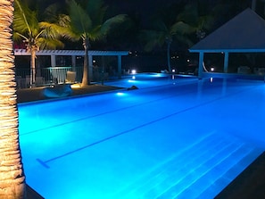 Une piscine éclairée de lumières bleues, entourée de palmiers et dotée d’un espace abrité.