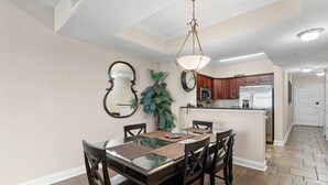 Un coin repas avec une table à plateau de verre, des chaises en bois, un luminaire suspendu, une horloge et, au fond, une cuisine.