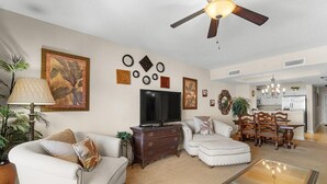 A living room with a ceiling fan, a TV, a glass coffee table, and a dining area with a chandelier. - Sun Sand Seclusion 2BR Oceanfront Condo Jetted Tub (North Myrtle Beach)