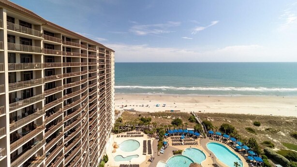 A high-rise hotel with multiple balconies overlooking a beach and a pool area with sun loungers and umbrellas. - Beachfront Elegance: 2BR Condo with Jetted Tub (North Myrtle Beach)