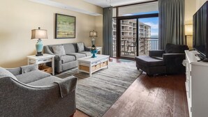 A modern living room with a grey sofa, a coffee table, and a TV. There is a balcony with a view of a building. - Beachfront Elegance: 2BR Condo with Jetted Tub (North Myrtle Beach)
