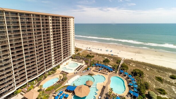 A high-rise hotel with multiple balconies overlooking a beach and a pool area with umbrellas and lounge chairs. - Spacious Coastal Comfort 2BR Condo with Balcony (North Myrtle Beach)