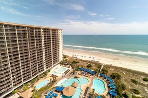 Ein Hochhotel mit mehreren Balkonen mit Blick auf einen Strand und ein Poolbereich mit Sonnenschirmen und Liegestühlen.