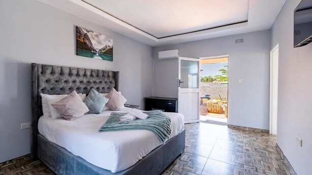 A bedroom with a large bed, a headboard, a painting on the wall, and a view of the outside. - The Crescent Victoria Falls  (Victoria Falls)