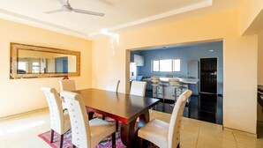 A dining area with a large table and chairs, a ceiling fan, and a kitchen visible through an archway. - The Crescent Victoria Falls  (Victoria Falls)