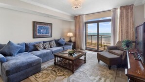 A living room with a sofa, armchair, coffee table, and a view of the ocean through a sliding glass door.