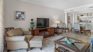 A living room with a sofa, ottoman, TV, and a glass coffee table.