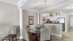 A dining area with a glass-top table and striped chairs, a kitchen with white cabinets, and a hanging chandelier.