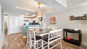 A dining area with a wooden table and white chairs, a kitchen in the background, and a wall-mounted word "Beach" sign.