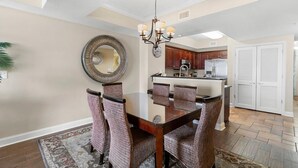 A dining area with a glass table and wicker chairs, a chandelier, and a kitchen in the background. - Penthouse Elegance by the Sea 3BR Condo with Views (North Myrtle Beach)