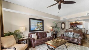 A living room with a brown leather sofa, a wooden coffee table, and a painting of a coastal scene. - Penthouse Elegance by the Sea 3BR Condo with Views (North Myrtle Beach)