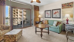A hotel room with a balcony, a green sofa, a round wooden table, and two lamps.