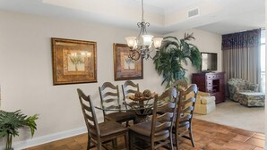 A dining area with a glass-top table, wooden chairs, and a chandelier. There are framed pictures on the wall, a large plant, and a television in the background.