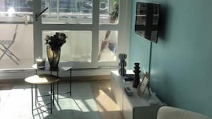 A modern living room with a large window, a flat-screen TV mounted on the wall, a white sofa, a small round table, and a potted plant. - sun and boat (Courseulles-sur-Mer)