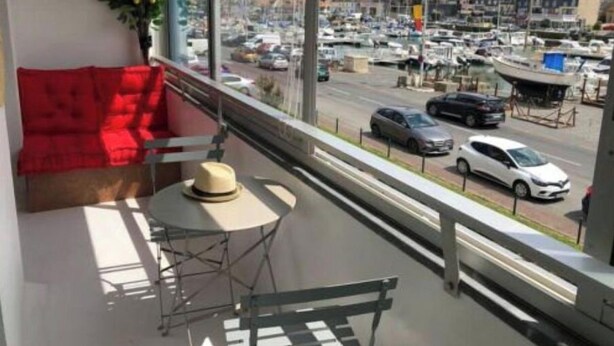 A balcony with a red sofa, a small round table, and a hat. A view of a marina with boats and cars is visible through the window. - sun and boat (Courseulles-sur-Mer)