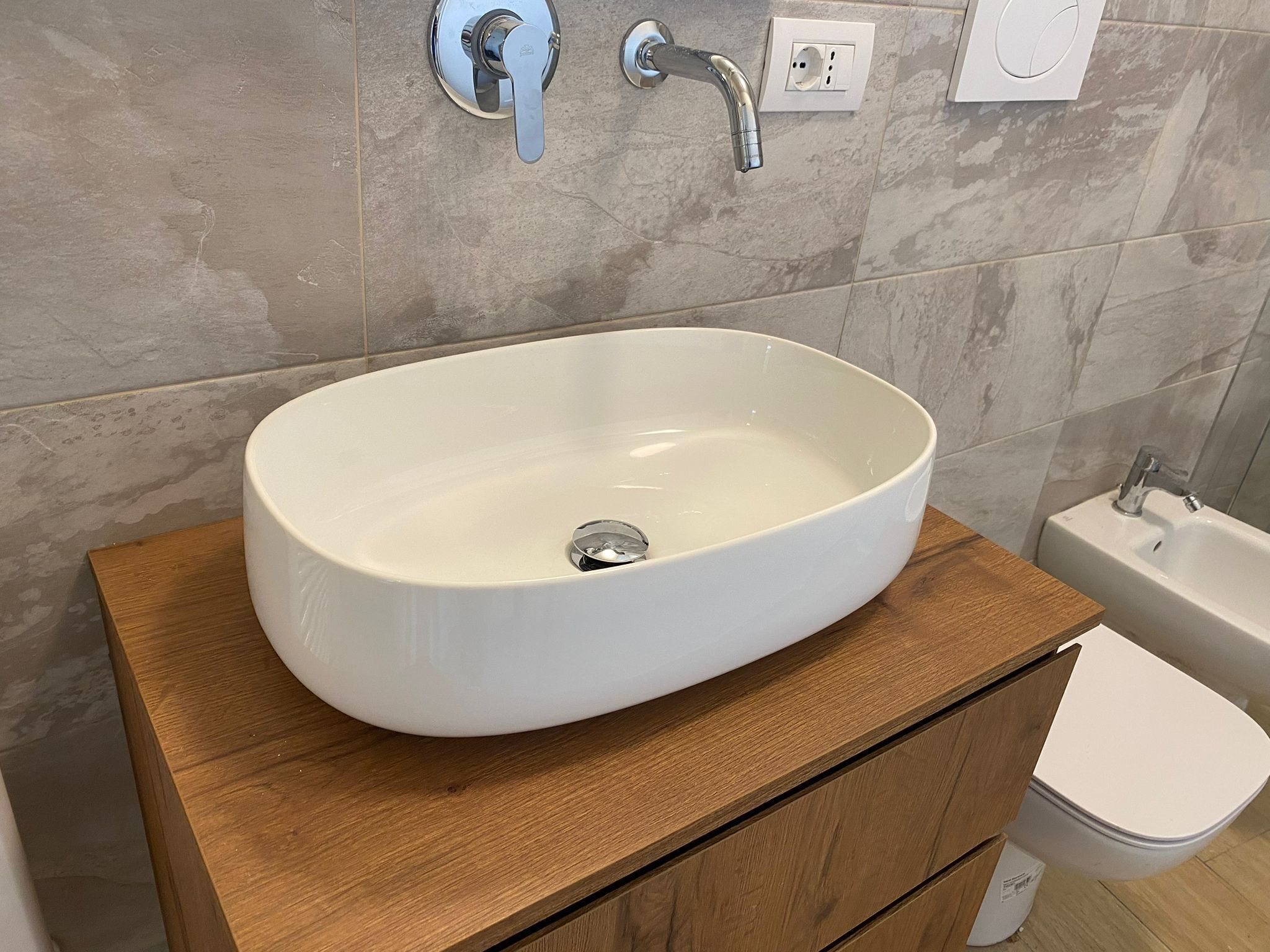 Une salle de bain avec un lavabo ovale blanc posé sur un meuble-lavabo en bois, des toilettes blanches et un espace douche.