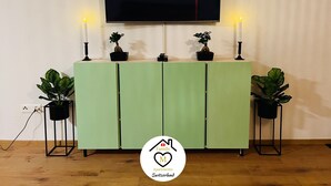 Ein Wohnzimmer mit einem grünen Schrank, zwei Blumentöpfen, einem Fernseher, der 'NETFLIX' anzeigt, und dem Logo von 'Family Apartments Switzerland'.